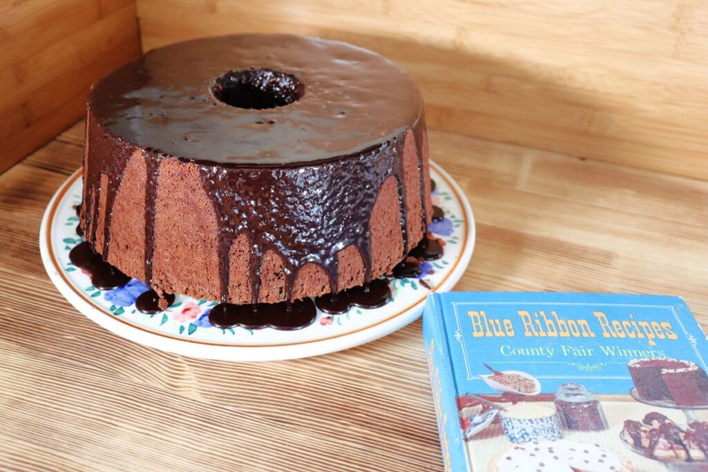 A chocolate glazed chocolate chiffon cake sits on a cake plate. Next to it is a copy of the book, 'Blue Ribbon Recipes.'