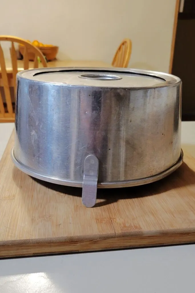 An aluminum tube pan sits upside down on its feet on a cutting board. A kitchen table and chairs in the background. 