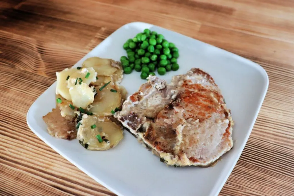 A pork chop sits on a square plate with potato slices garnished with snipped chives and some green peas. 