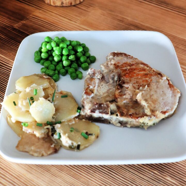 A pork chop sits on a square plate with sliced potatoes and green peas.