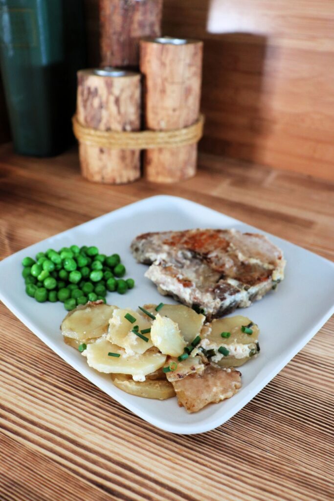 Slices of potatoes garnished with chives sit at the front of a plate with a pork chop and peas behind them. In the background are some candles and a book. 