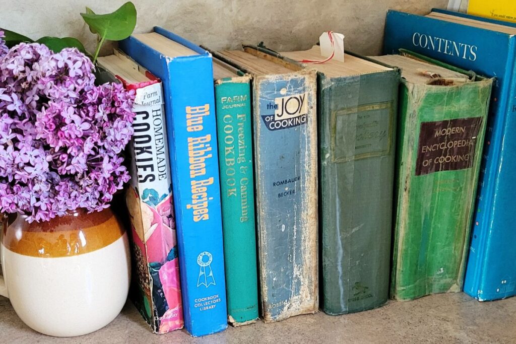 A shelf full of vintage cookbooks held in place by a vace of lilacs.