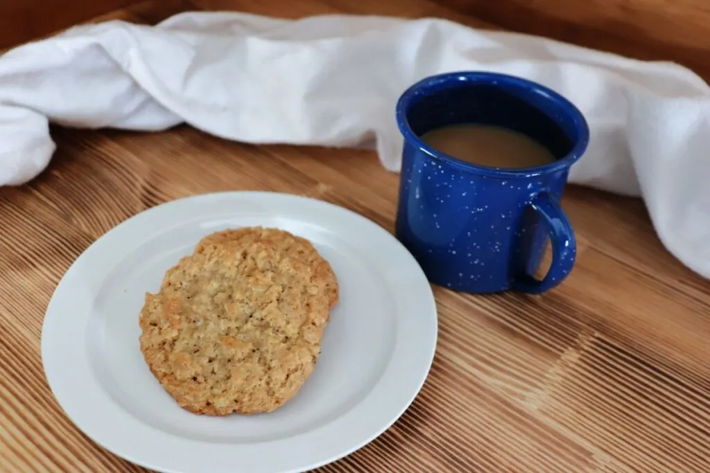 2 cookies sit on a white plate next to a blue cup with coffee inside, a white cloth draped behind everything. 