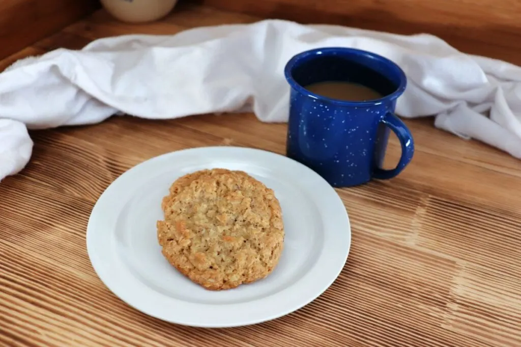 2 cookies sit on a white plate next to a blue cup with coffee inside, a white cloth draped behind everything. 