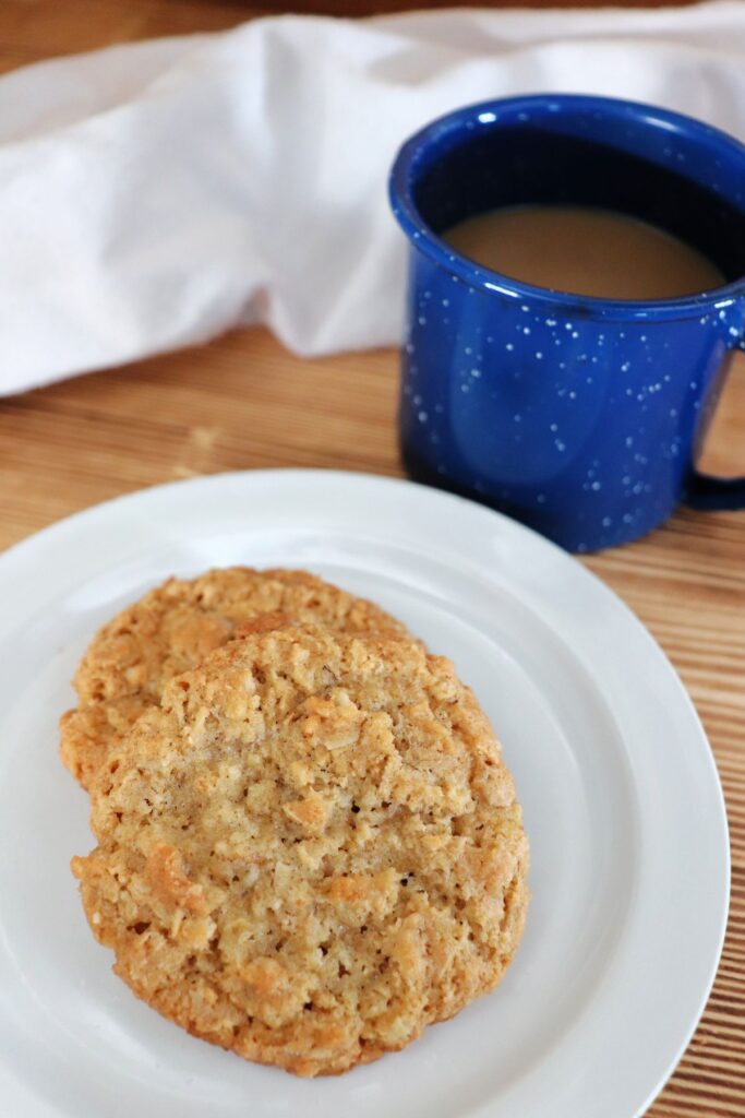 A close-up of 2 cookies sitting on a white plate next to a blue cup with coffee inside, a white cloth draped behind everything. 