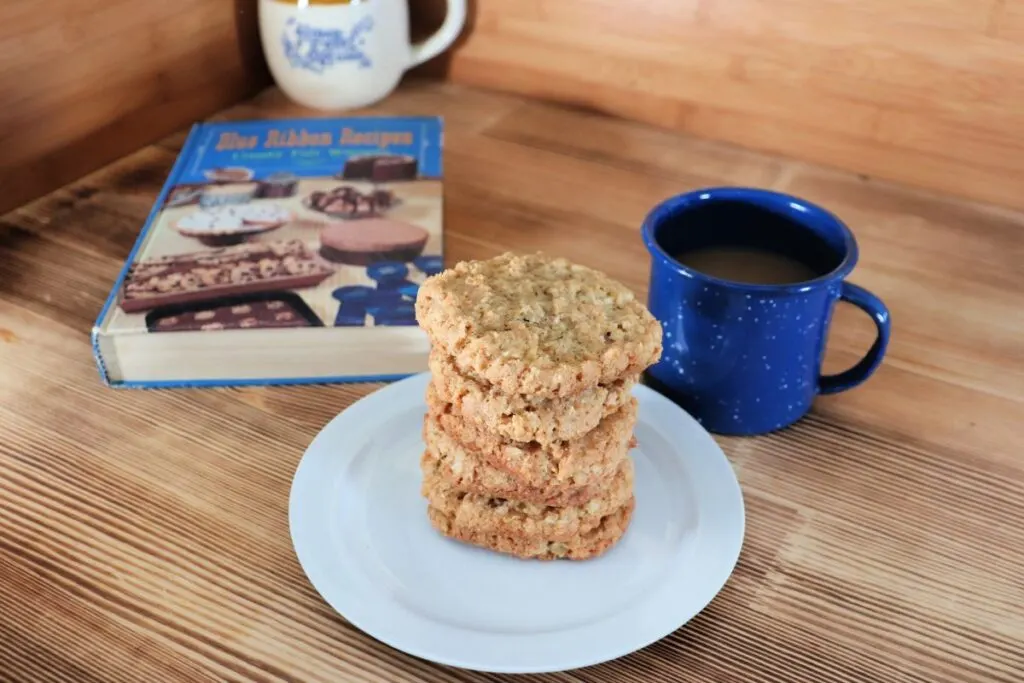  Cookies are stacked on a white plate with a blue cup of coffee next to it. Behind the plate sits a vintage cookbook, titled 'Blue Ribbon Recipes'.