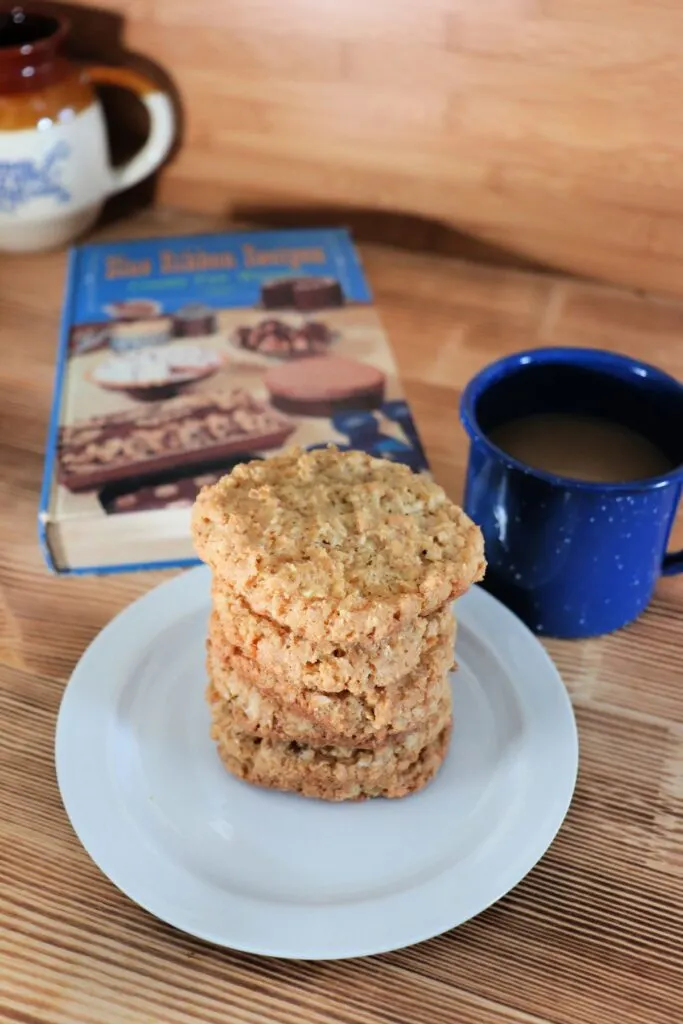  Cookies are stacked on a white plate with a blue cup of coffee next to it. Behind the plate sits a vintage cookbook, titled 'Blue Ribbon Recipes'.