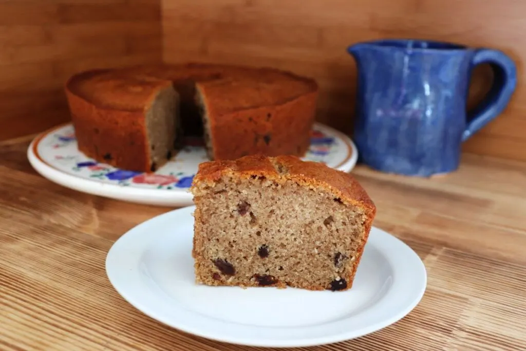 A slice of raisin spice cake sits on a white plate with the remaining cake in the background.