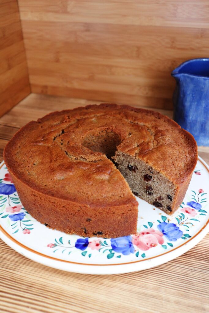 A raisin spice cake sits on a floral cake plate with a slice removed from it. A blue pitcher sits in the background.