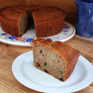 A slice of raisin spice cake sits on a white plate with the remaining cake in the background.