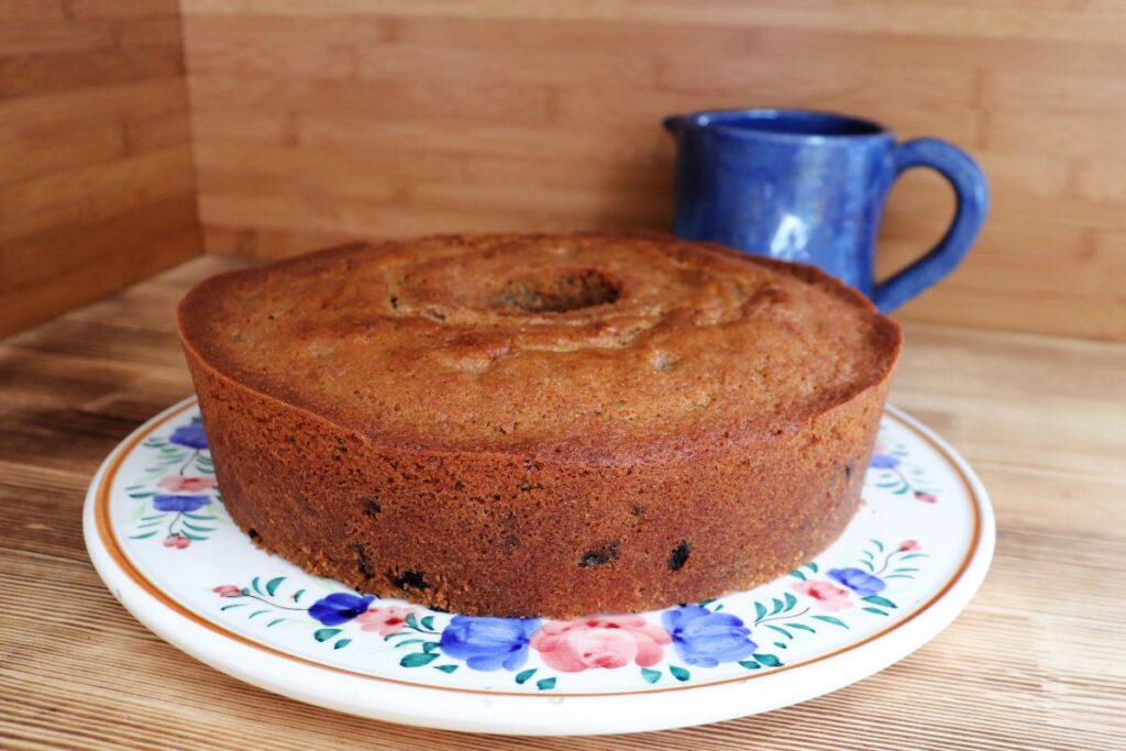 A whole raisin spice cake sits on a floral cake plate with a blue pitcher in the background.