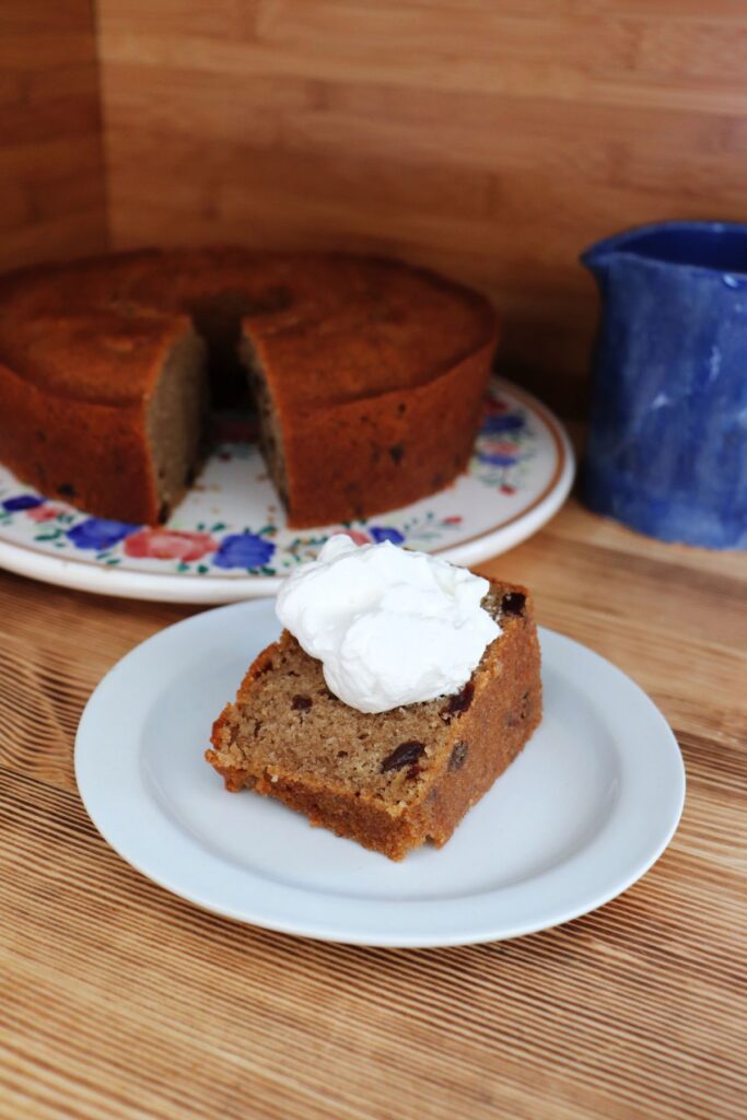 A slice of raisin spice cake with whipped cream sits on a plate with the remaining cake and a blue pitcher in the background.