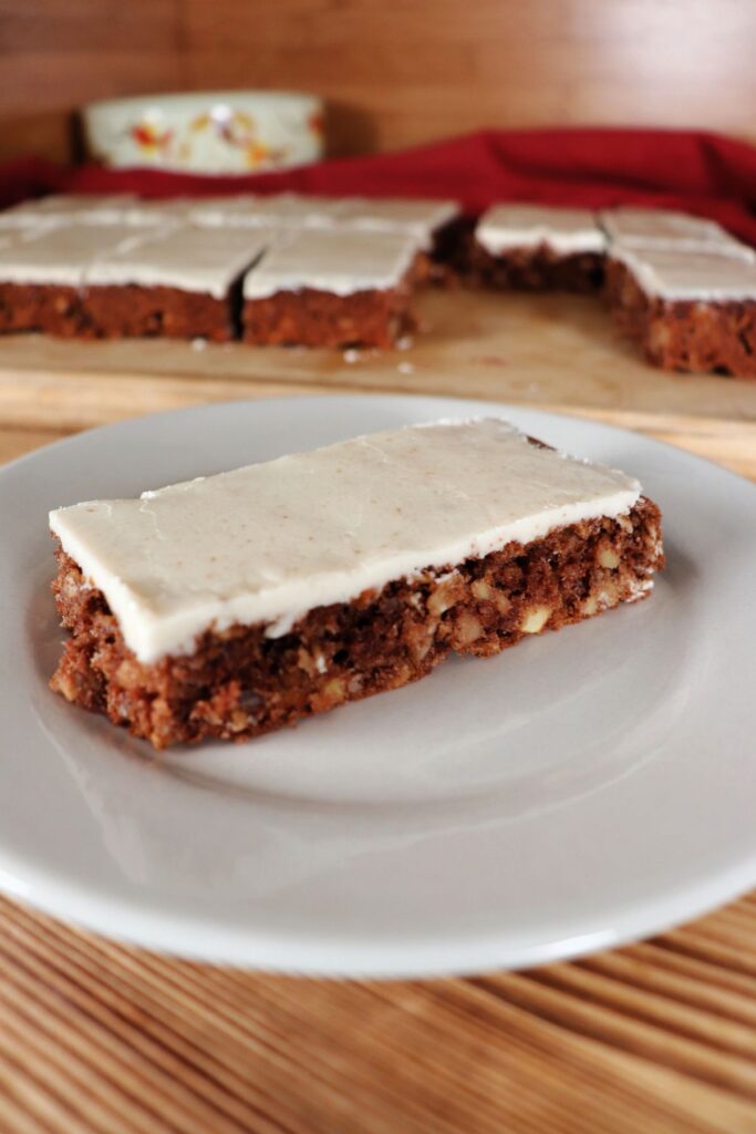 A caramel frosted apple oat bar as seen from the side sits on a white plate. In the background sits a board with more bars on it. 