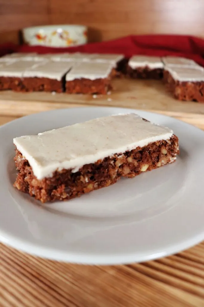 A caramel frosted apple oat bar as seen from the side sits on a white plate. In the background sits a board with more bars on it. 