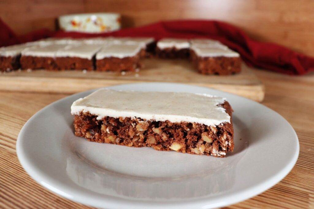 A caramel frosted apple oat bar as seen from the side sits on a white plate. In the background sits a board with more bars on it. 