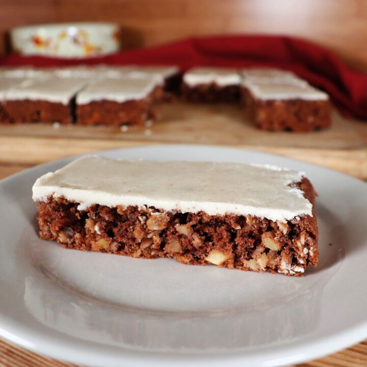 A caramel frosted apple oat bar sits on a white plate with a platter of more bars in the background.