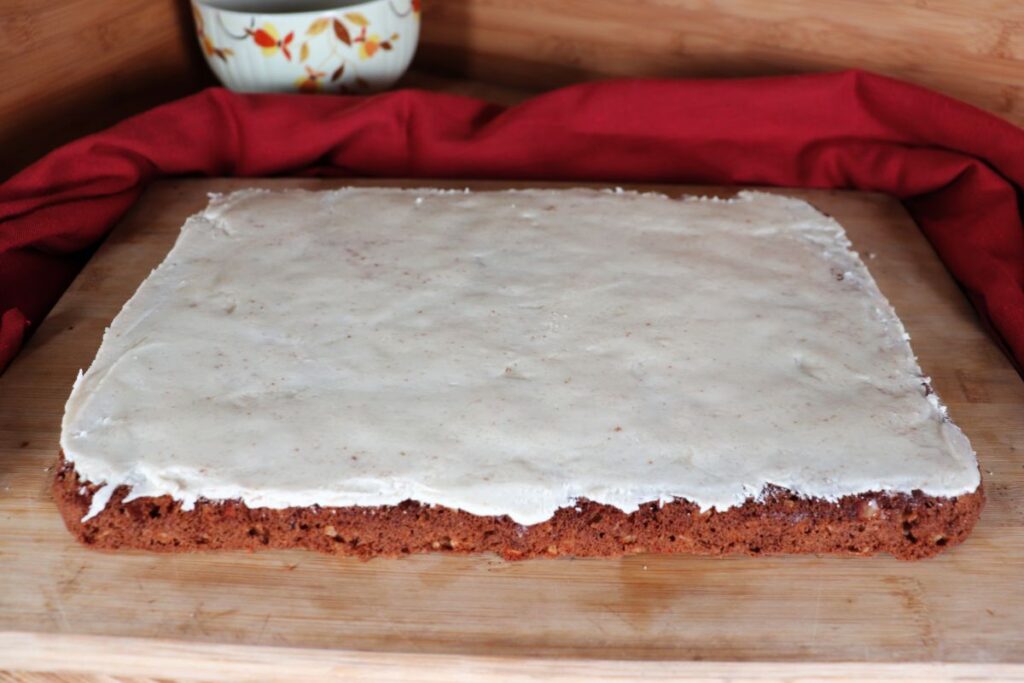 An uncut pan of frosted apple oat bars sits on a wooden cutting board waiting to be sliced. The board is surrounded by a red cloth. 