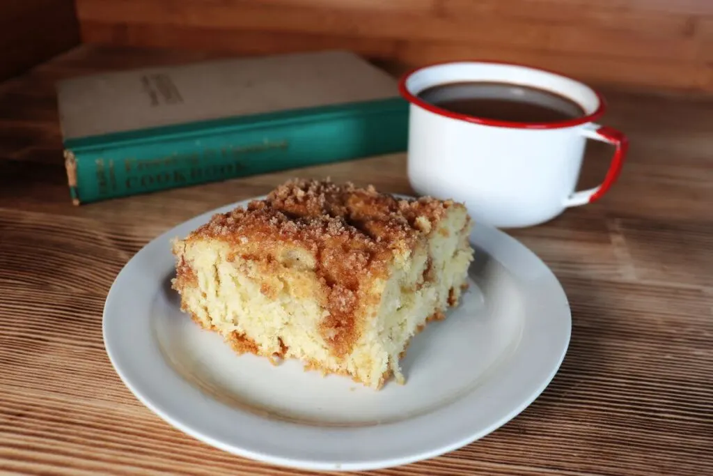 A slice of streusel topped coffee cake sits on white plate with a coffee cup and book behind it. 