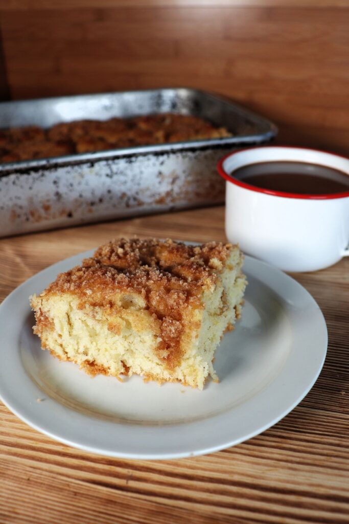 A slice of streusel topped coffee cake sits on white plate with a coffee cup and remaining cake in a pan behind it. 