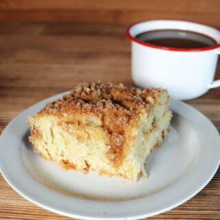 A slice of coffee cake sits on a white plate. A white cup with a red rim full of coffee sits behind it.