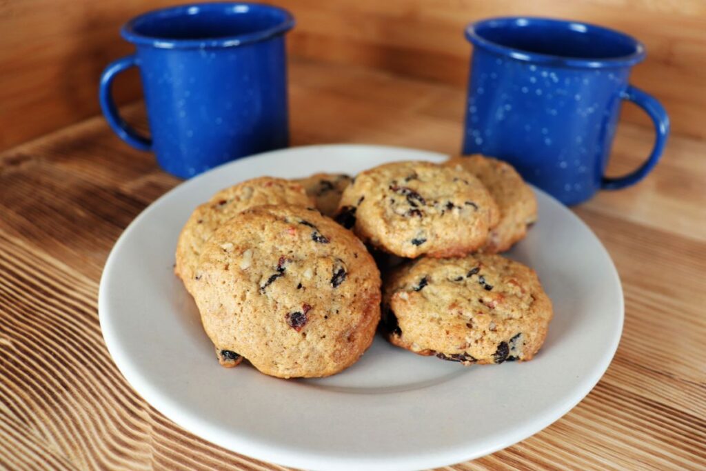 Cookies showing bits of nuts and dried fruits are stacked on a plate with blue coffee cups behind it.