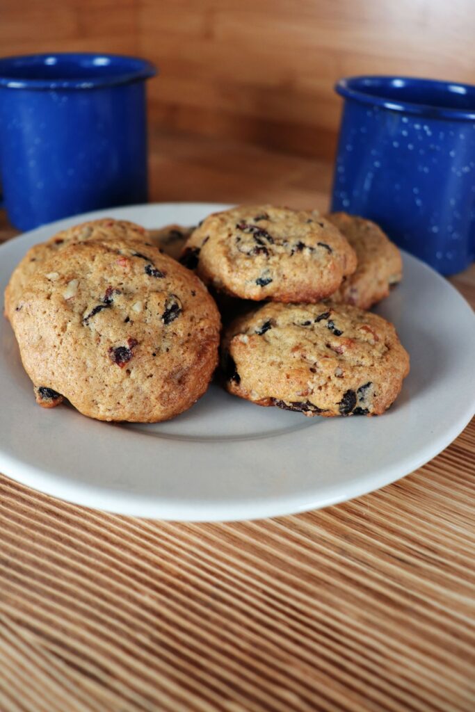 Cookies showing bits of nuts and dried fruits are stacked on a plate with blue coffee cups behind it.