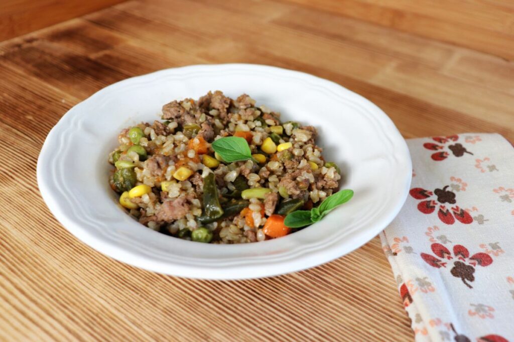 A white bowl of a hamburger rice casserole sits on a table. The meal is garnished with fresh herbs and shows rice, meat, and veggies.